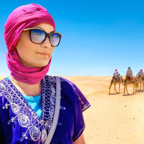 Portrait of beautiful woman in arabic traditional clothing against background of tourists riding on camels. Sahara desert, Tunisia, North Africa